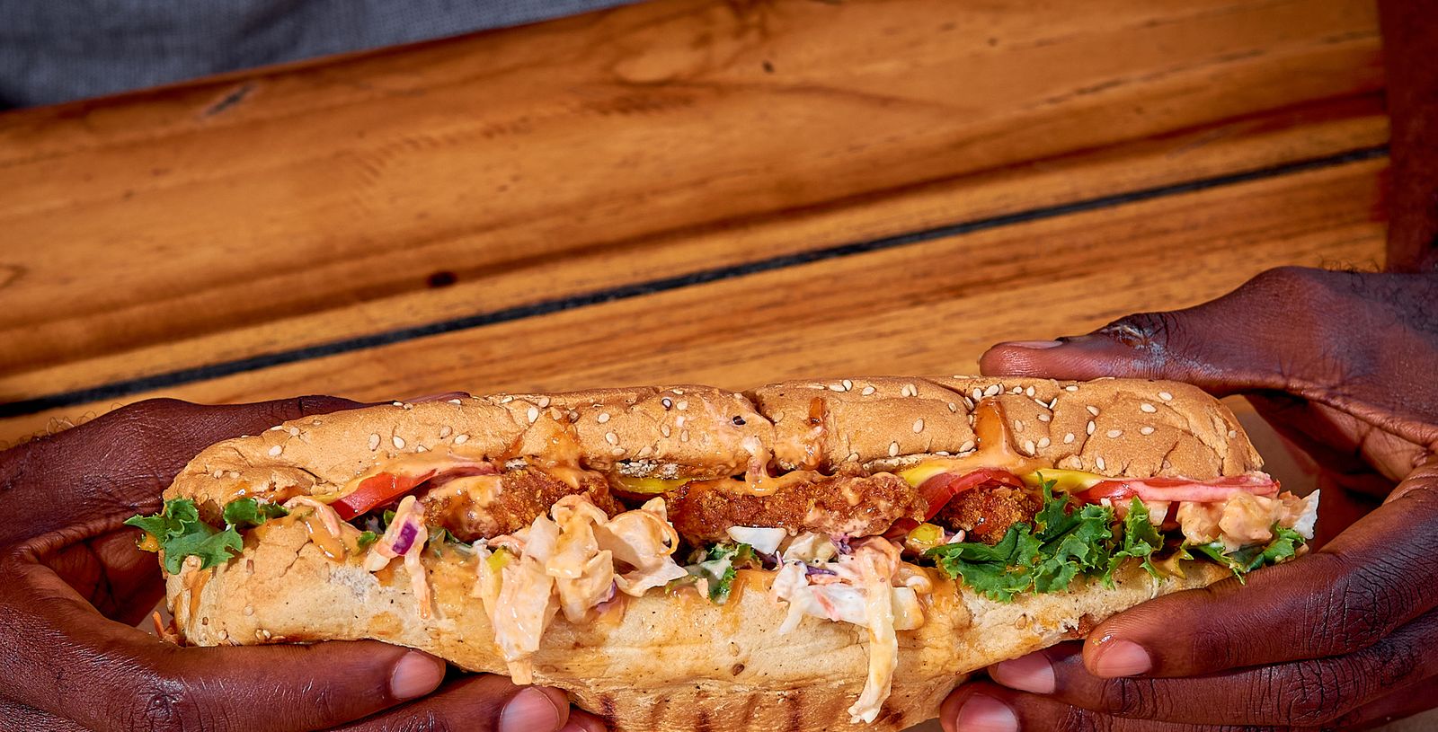 A freshly built sub held in both hands over a wooden counter